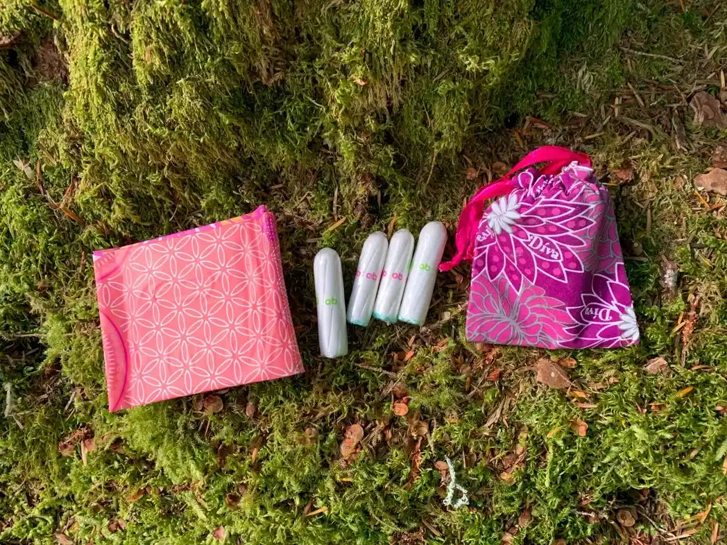 Menstrual products including pads, tampons and a cup rest on a mossy forest floor. Learn how to camp and hike on your period