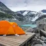 An orange tent sits on a wooden tent platform in front of a blue lake and a mountain with glaciers. Backcountry camping at Wedgemount Lake in Garibaldi Provincial Park.