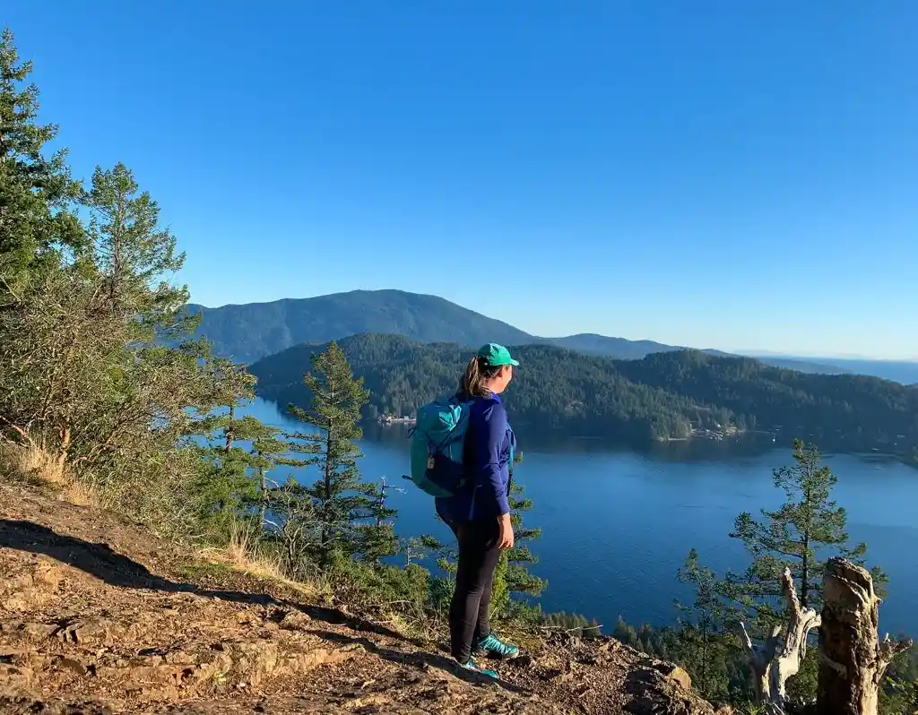 A female hiker wearing hiking leggings and a backpack stands on top of Soames Hill looking down at the ocean