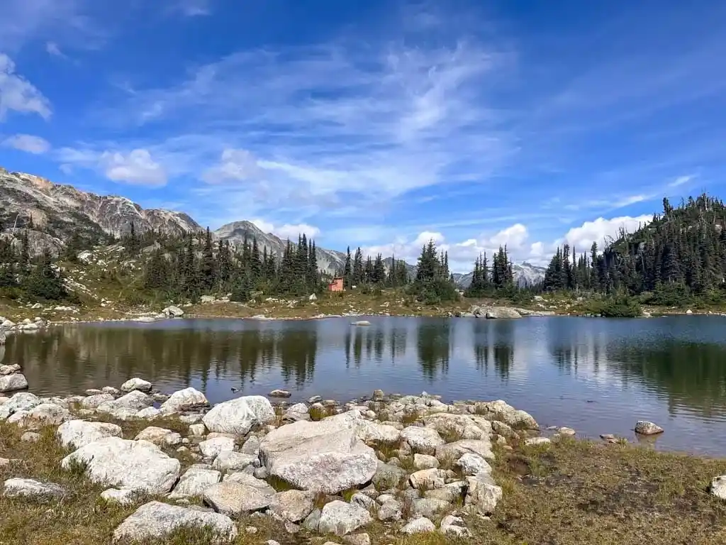 An alpine lake with rocks in the foreground. You can see a small wooden outhouse building on the other side of the lake.
