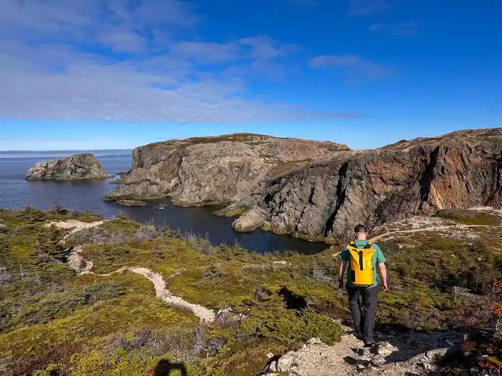 A man in a yellow backpack walks along a coastal trail near headlands on the Rockcut Trails in Twillingate