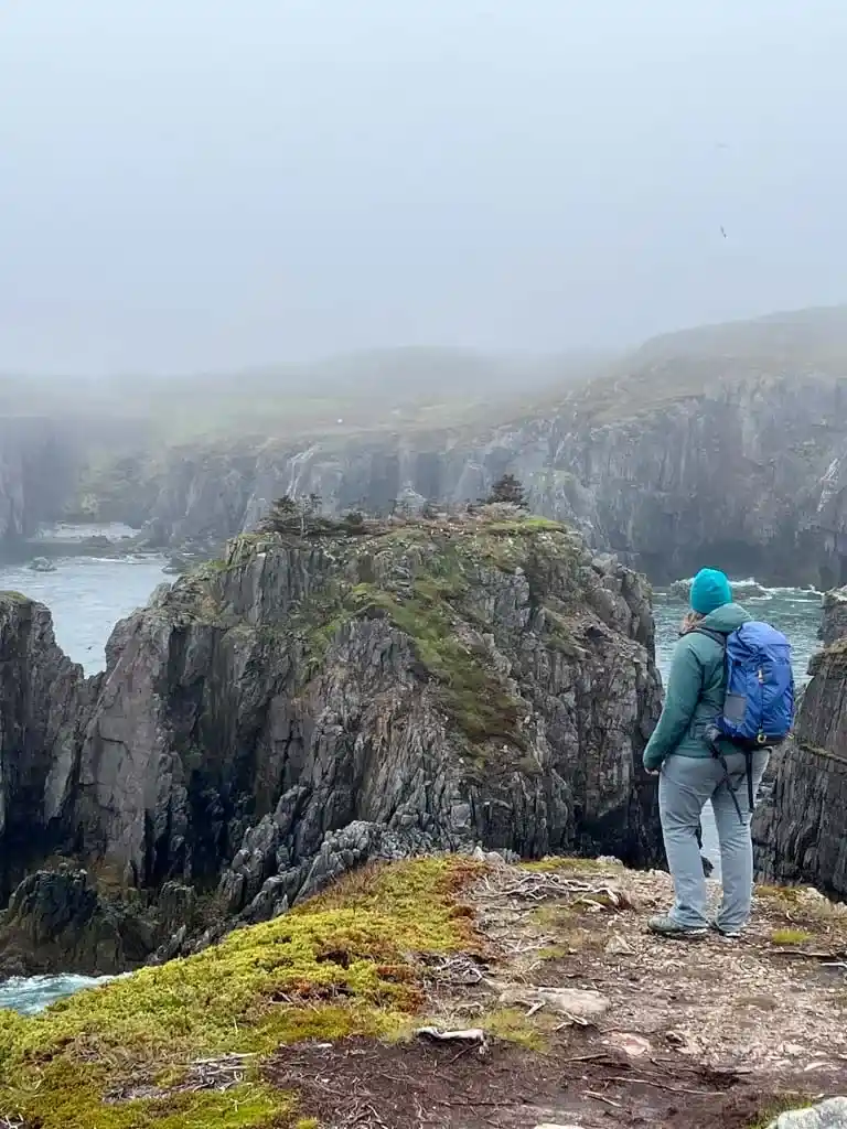 A woman stands at the edge of a cliff watching puffs on an offshore island near Bonavista Newfoundland. She is dressed for hiking with a backpack, warm jacket, and fleece hat.